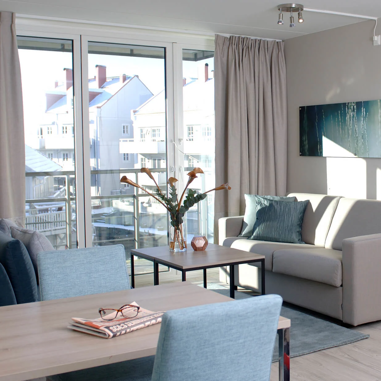 Stue med beige sofa og turkise puter, foran et spisebord med avis og briller. Store vinduer med utsikt til nabobygninger, koblet til soverom med turkist sengetøy.