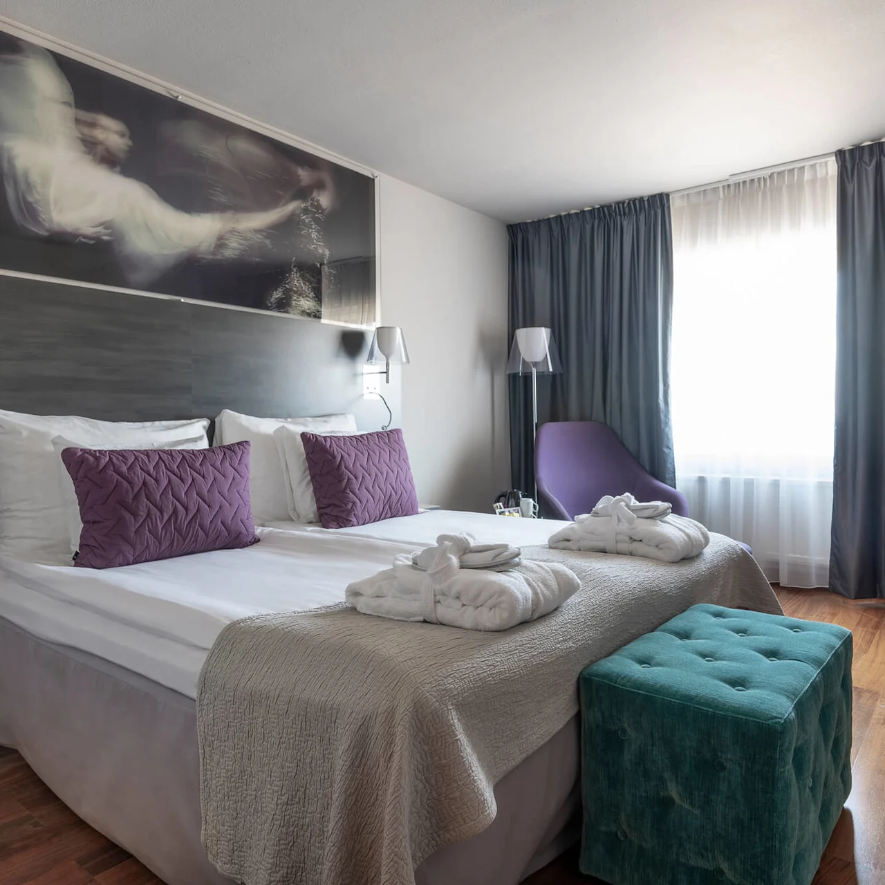Ett prydligt arrangerat hotellrum med en dubbelsäng med två lila kuddar och vita morgonrockar. En turkos ottoman står vid fotändan, med ett skrivbord och stol i närheten.