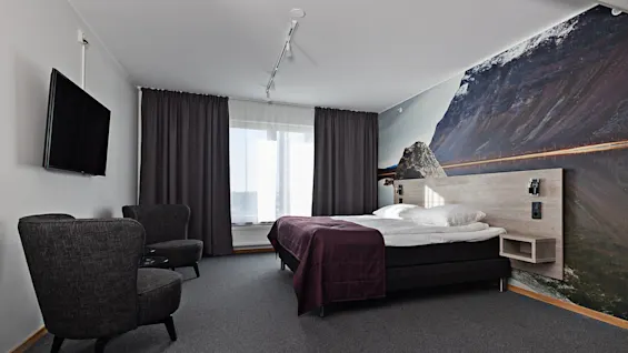 Ett modernt hotellrum har en dubbelsäng med ett lila överkast, två grå fåtöljer riktade mot en väggmonterad platt-TV och en naturskön bergsmålning på en vägg.