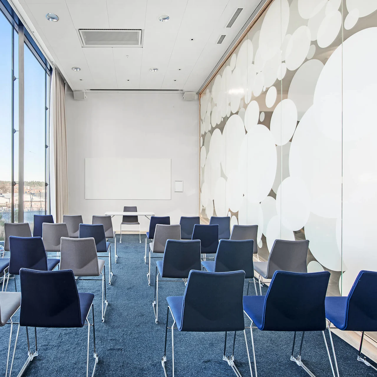 Rækker af blå og grå stole vender mod en tavle i et moderne konferencelokale med et stort vindue med udsigt over en marina og en dekorativ væg med cirkulære mønstre.