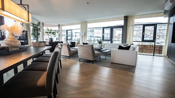 Romslig stue med et langt spisebord og stoler, moderne sofaer og lenestoler. Store vinduer slipper inn naturlig lys og gir utsikt over byens bygninger utenfor.