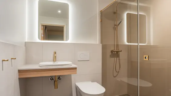 Ett modernt badrum med handfat med guldkran, toalett och dusch med glasvägg. Bakgrundsbelyst spegel och varm belysning.