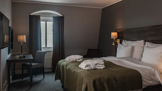 A neatly made bed with pillows and robes lies against a wall. A desk with a chair is near a window with closed curtains, creating a cozy hotel room ambiance.