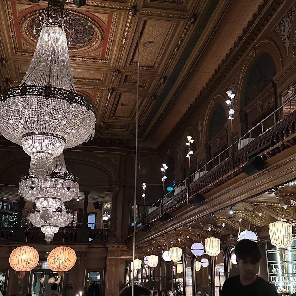 En storslået, udsmykket sal med funklende lysekroner og dekorative lofter, fyldt med mennesker, der nyder en aften ude.