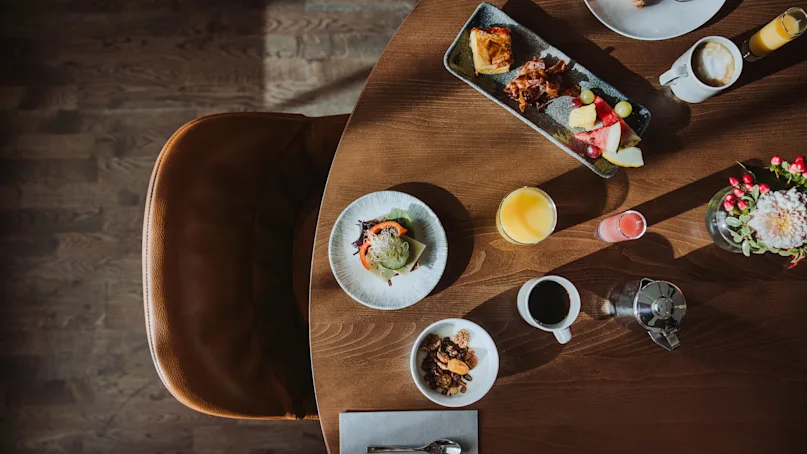 A round wooden table holds a diverse breakfast spread, including a sandwich, juice, coffee, and fruit with nearby flowers, set in a sunny dining room.