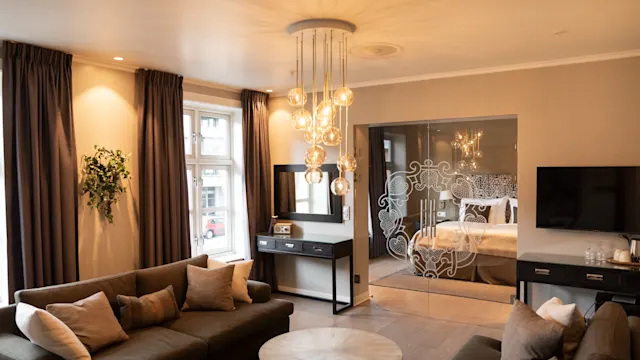 Et moderne oppholdsrom med to brune sofaer vendt mot en dekorativ glassvegg. Myk belysning fra en lysekrone lyser opp rommet, med store vinduer og mørke gardiner.