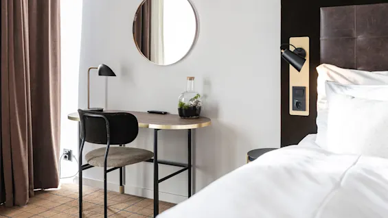 Skrivebordet har en lampe, fjernbetjening og terrarium; placeret ved siden af en seng med brunt hovedgærde. Et rundt spejl hænger over i et rum med brune gardiner og tæppe.