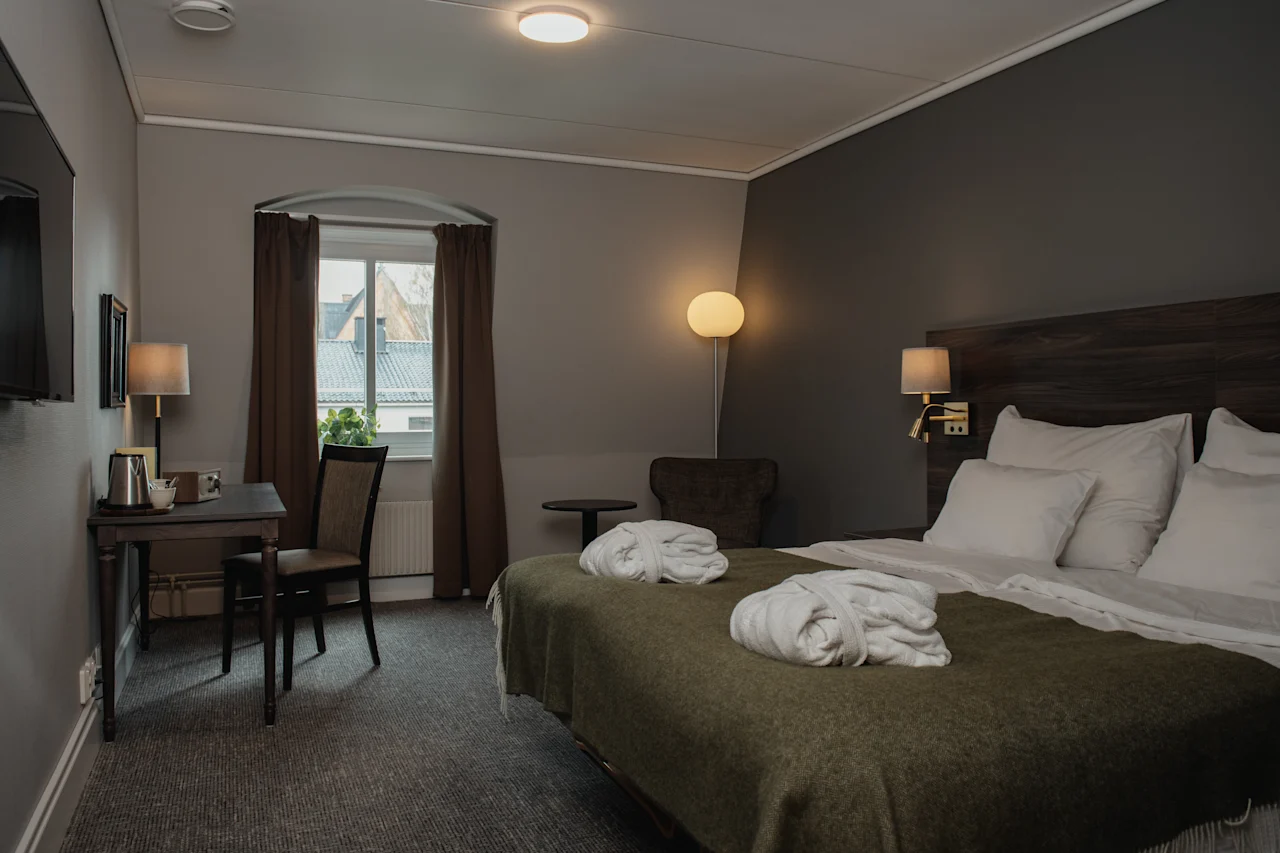 A bed adorned with white robes rests in a warmly lit hotel room, featuring a desk, chair, window with curtains, and a floor lamp in a cozy atmosphere.