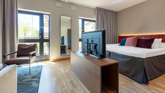 A modern bedroom with a large bed, armchair, television, and balcony access in a bright, airy space.