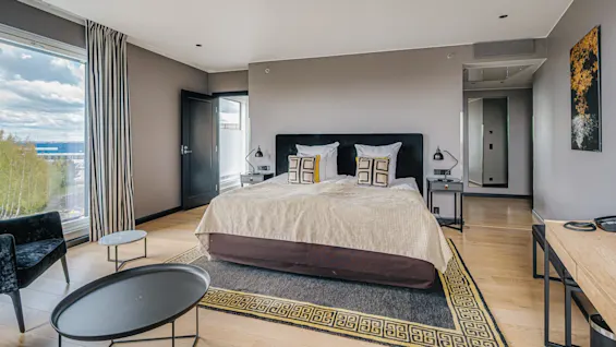 A bed with decorative pillows is centered in a minimalist hotel room, featuring wooden floors and a large window with a view of trees and clouds.