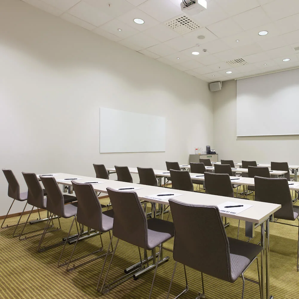 Classroom setting with empty chairs and desks arranged in rows facing a large projection screen. Notepads and pens are placed neatly on each desk. The room features whiteboards and overhead lighting.