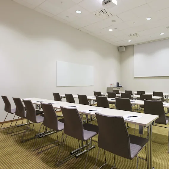 Klassrum med tomma stolar och skrivbord i rader mot en stor projektionsskärm. Anteckningsblock och pennor ligger prydligt på varje bord. Rummet har whiteboards och takbelysning.