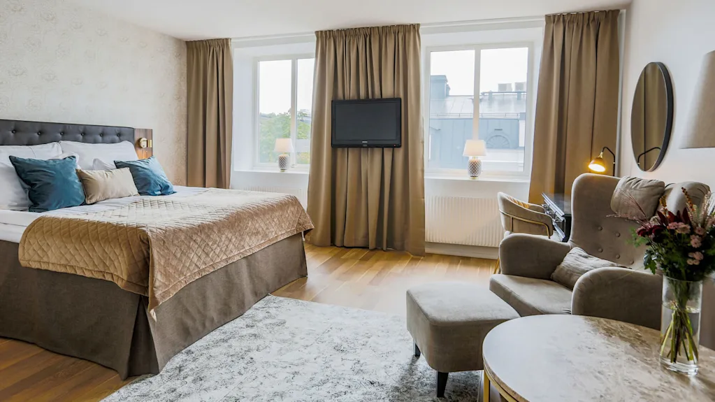 A neatly arranged bedroom with a bed, chairs, and a TV, showcasing a cozy interior with natural light from windows.