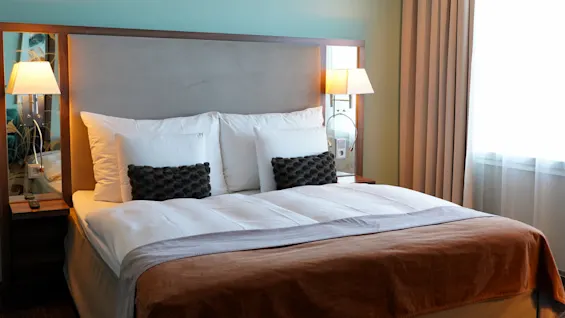 A neatly made bed with white and brown bedding, flanked by two bedside tables each holding a lit lamp, in a softly lit room with teal walls and beige curtains.