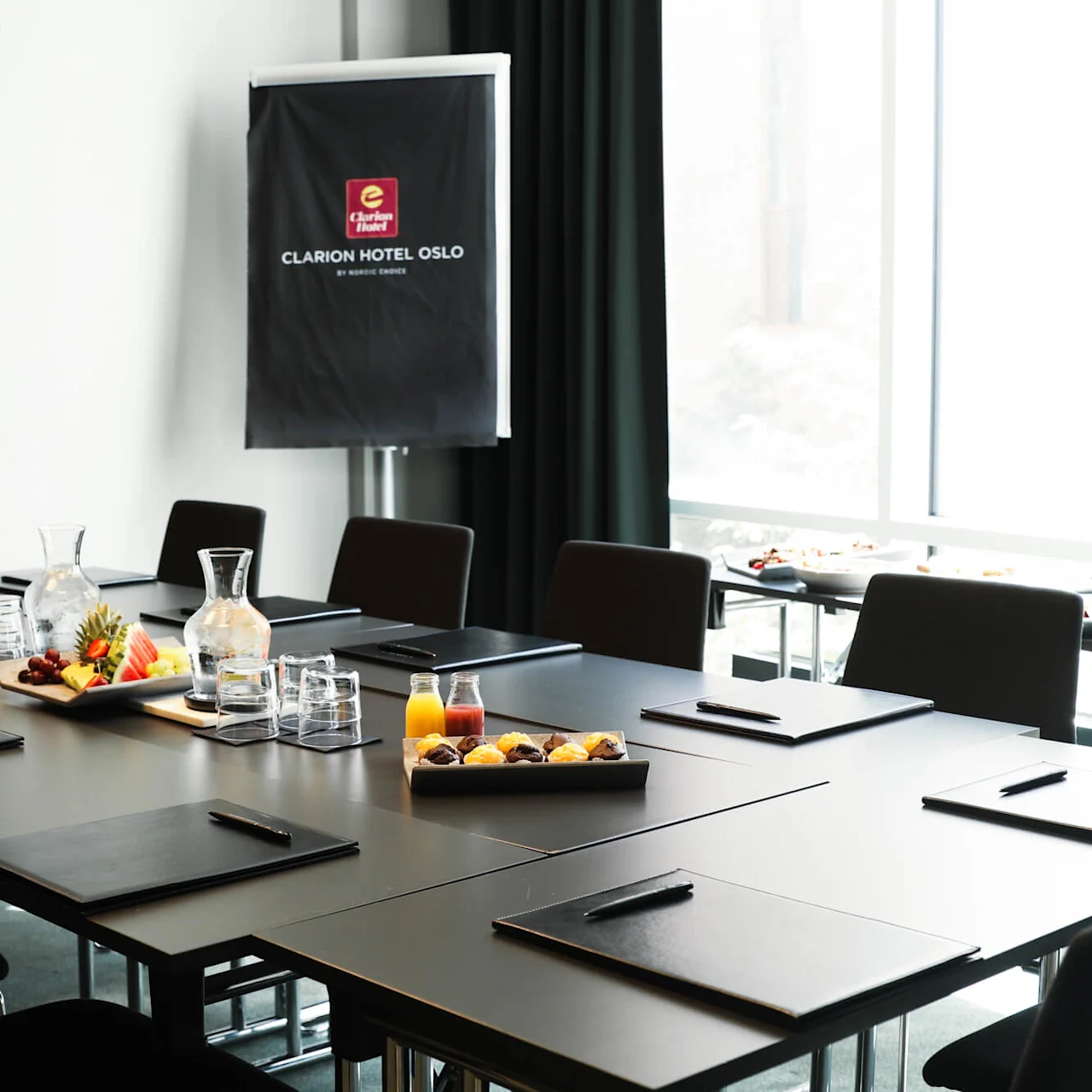 A conference room with arranged tables, folders, water carafes, fruit platters, and juices. "CLARION HOTEL OSLO BY NORDIC CHOICE" is displayed on a standing board near a screen.