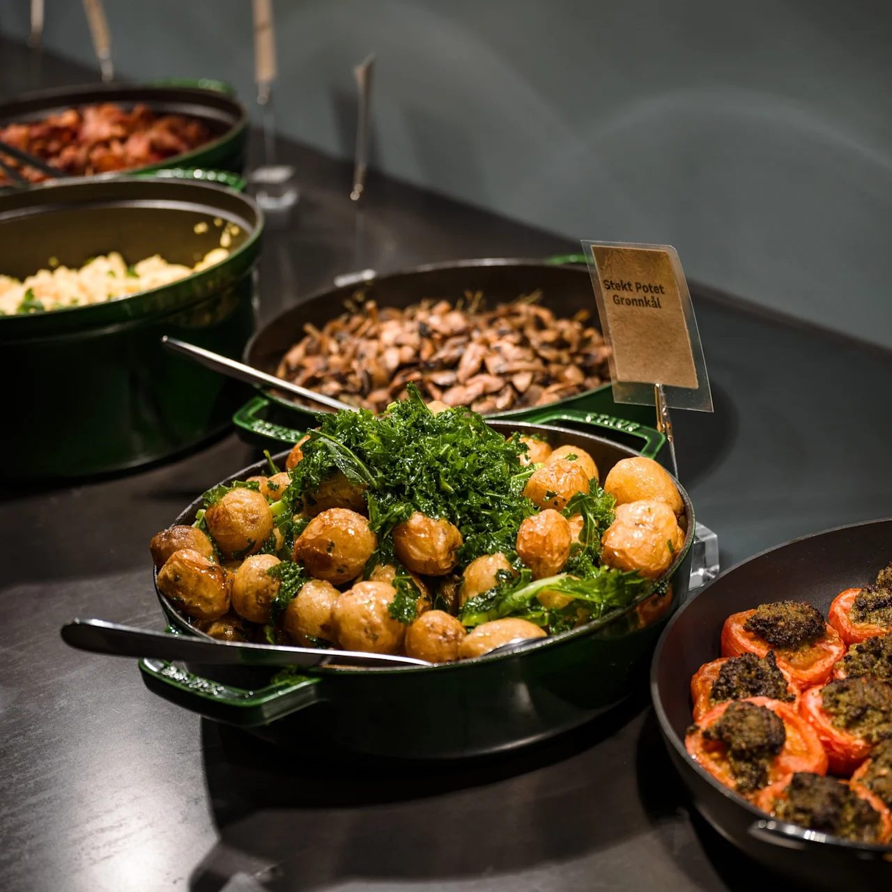 Gyldne ristede kartofler med grønkål anrettet i en grøn skål på et buffetbord, ledsaget af et skilt med teksten "Stekt Potet Grønnkål", omgivet af andre retter inklusiv tomater og svampe.