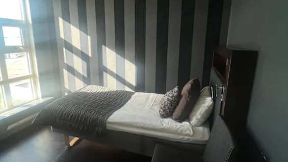 A single bed with pillows rests against a striped wall in a minimally furnished room, illuminated by sunlight streaming through a window on the left.