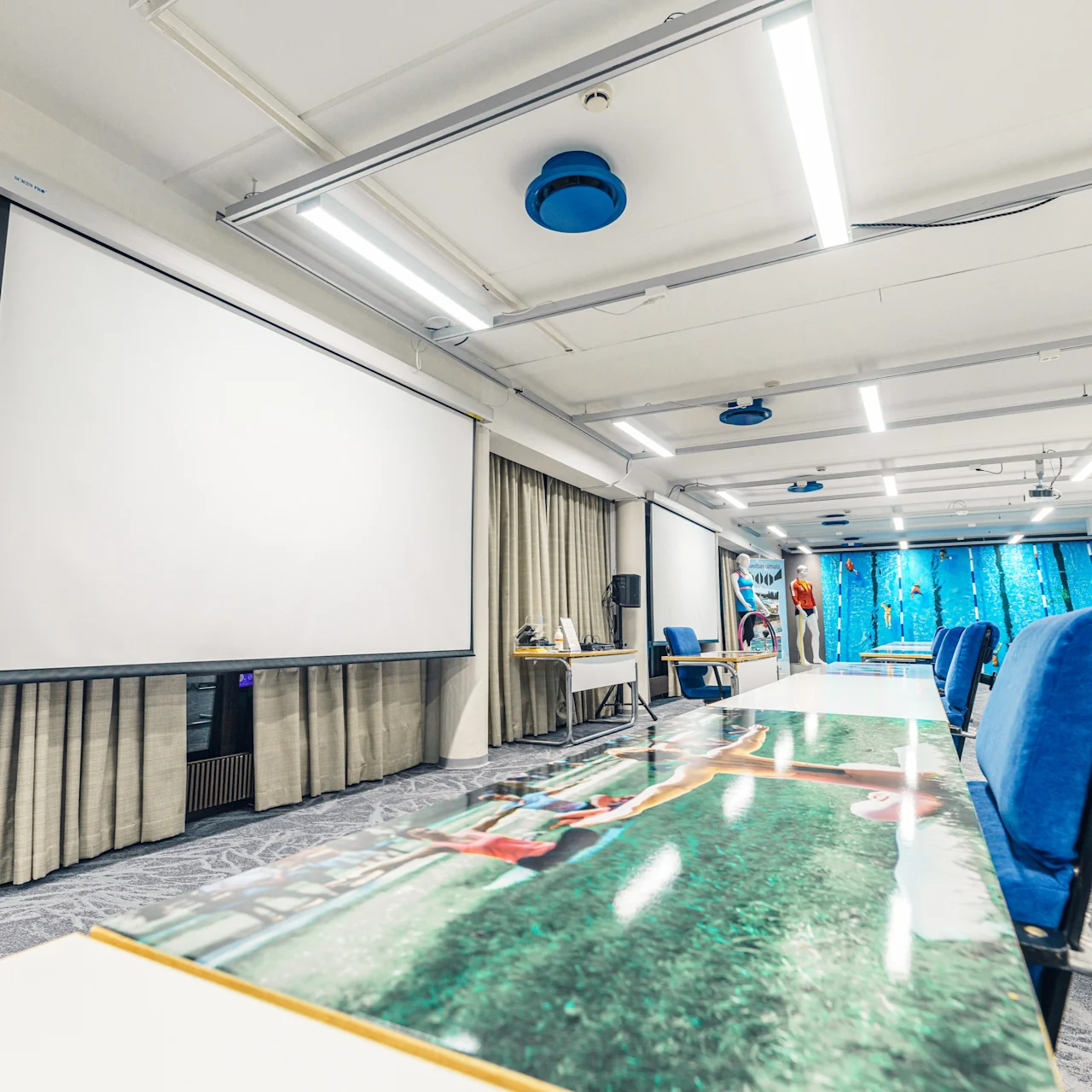A large conference room with blue chairs and a photo-printed table surface, facing a blank projection screen. Curtains line the walls, and overhead lights illuminate the space.