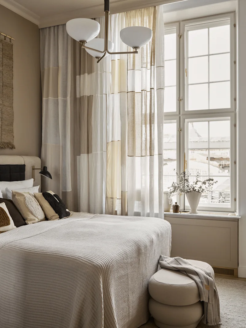 Seng pyntet med puder i et hyggeligt soveværelse. Naturligt lys filtreres gennem gardiner i patchwork design. Ved siden af, en vindueskarm med dekorative vaser forstærker den rolige atmosfære.
