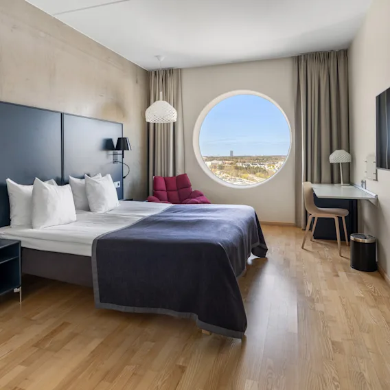 Modern hotel room with wooden floors, a large double bed, a round window with a view, and a wall-mounted TV.
