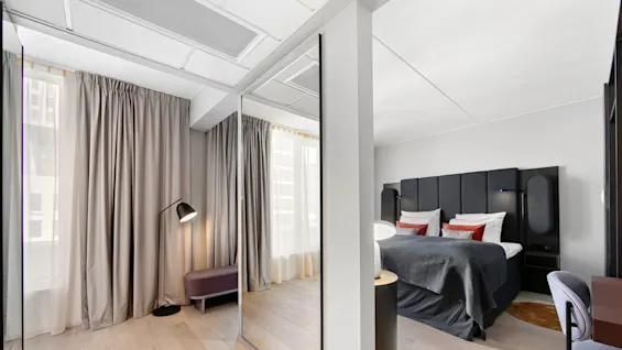 A modern hotel room features a bed with dark headboard and bedding, a bedside table with lamp, and a corner with floor lamp, reflected in a mirrored wardrobe.