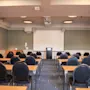 Et veloplyst konferencelokale med rækker af borde og stole vendt mod en stor projektorskærm og et podium.