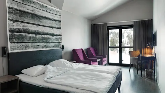 A bed with white linens sits in a modern room, accompanied by purple chairs and a desk with a lamp. Large windows reveal a snowy forest outside.