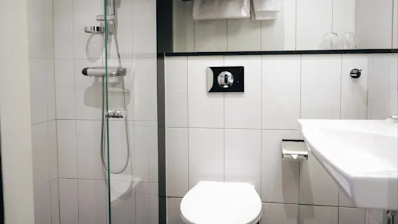Dusj med glassdør står i et hvitkaklet baderom. I nærheten er det et toalett med innebygd spyleknapp. To håndklær henger over toalettet, og en servant er synlig.