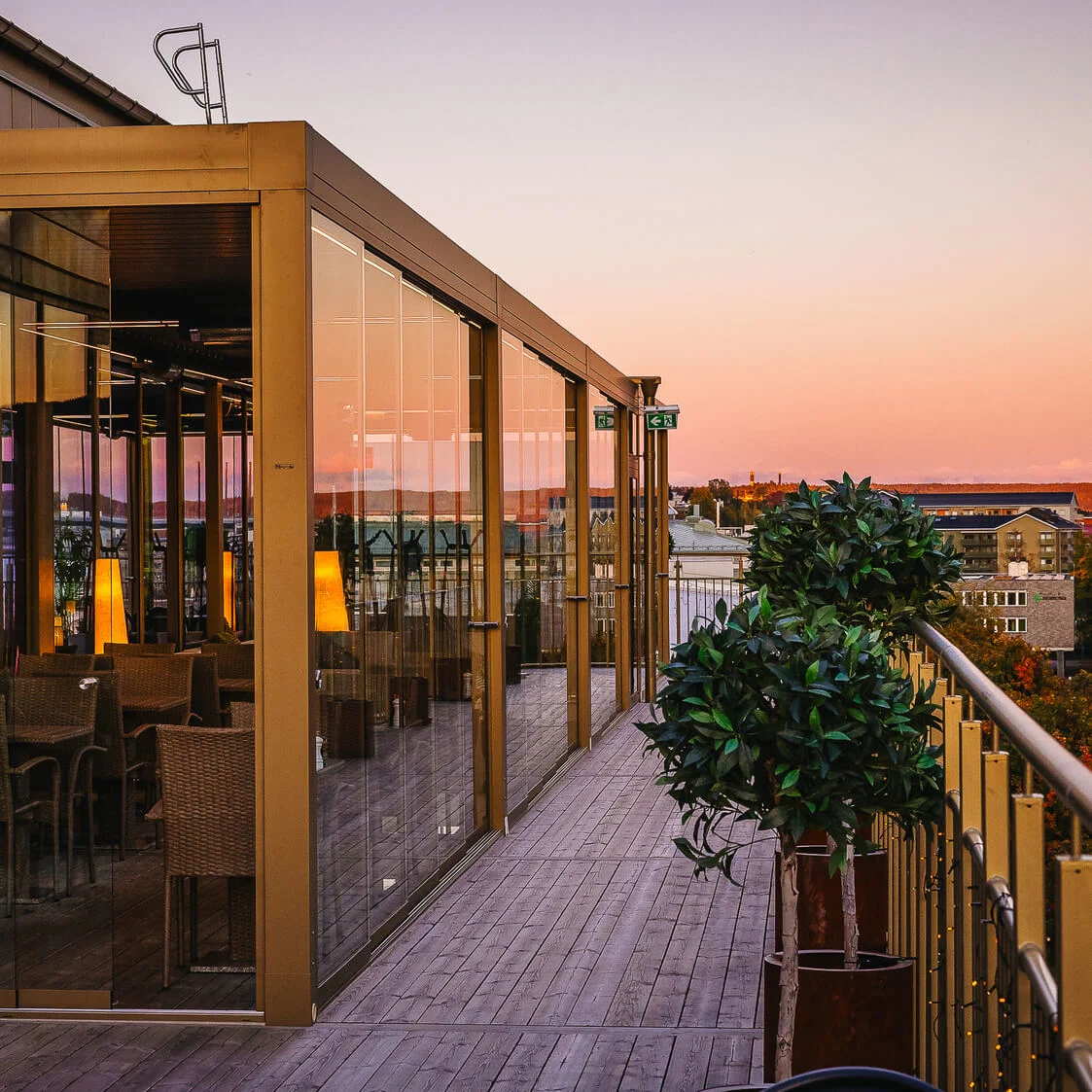 Glasindelukket terrasse med kurvestole indeni, reflekterer en solnedgang. Omgivet af byens bygninger og træer, fører en trægang langs rækværket med potteplanter.