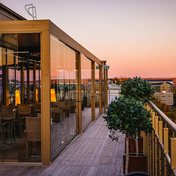 Glass-enclosed terrace with wicker chairs inside, reflecting a sunset. Surrounded by city buildings and trees, the wooden pathway adorned with potted plants leads along the railing.  