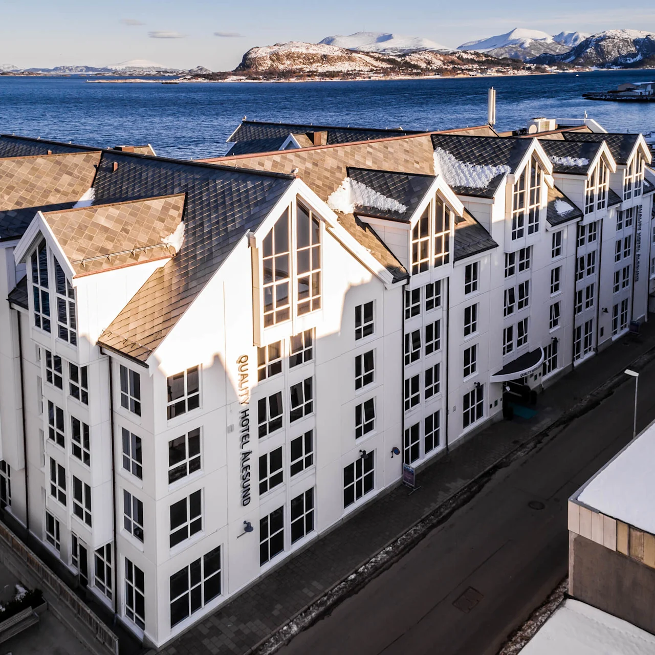 Et stort, hvidt hotel med stejle gavltag overser et blåt hav. Bjerge er synlige i baggrunden. Tekst på bygningen lyder "QUALITY HOTEL ÅLESUND."