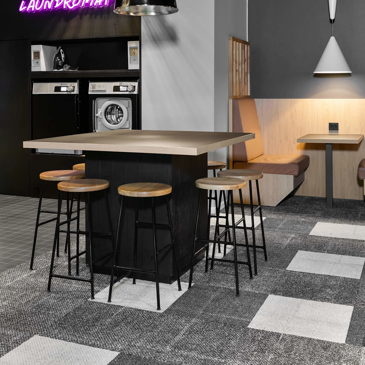 Washing machines beneath a neon "LAUNDROMAT" sign in a modern, stylish room with wooden bar stools around a table, grey walls, and patterned flooring.