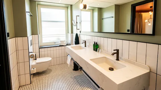 A modern bathroom features a double sink with white marble countertops, a wall-mounted toilet, and large mirror. Sparse decor includes stacked towels and soap dispensers, set against green and white tiling.