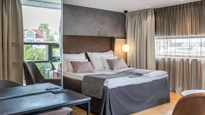 A neatly made bed with pillows and a brown headboard stands in a modern room, featuring soft lighting and large windows, with a desk and leather chairs nearby.