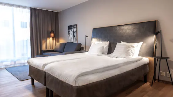 A modern bedroom features a neatly made double bed with a dark headboard, adjacent to a grey sofa and lamp. Curtains cover large windows, creating a cozy atmosphere.