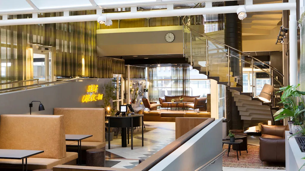 En elegant hotellobby med moderne møbler, et skilt for selvbetjening og en spiraltrappe, der fører op til et øvre niveau.