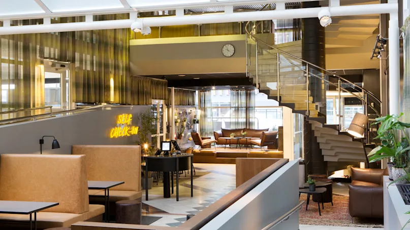 An elegant hotel lobby with modern furniture, a self check-in sign, and a spiral staircase leading to an upper level.
