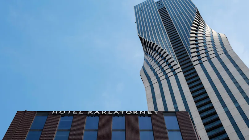 A twisting skyscraper towers upward against a clear blue sky. Below, a modern building facade displays the text "HOTEL KARLATORNET".