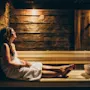 En kvinde, indhyllet i et håndklæde, slapper af på en træbænk i en svagt oplyst sauna, omgivet af rustikke trævægge, med en træspand i nærheden.