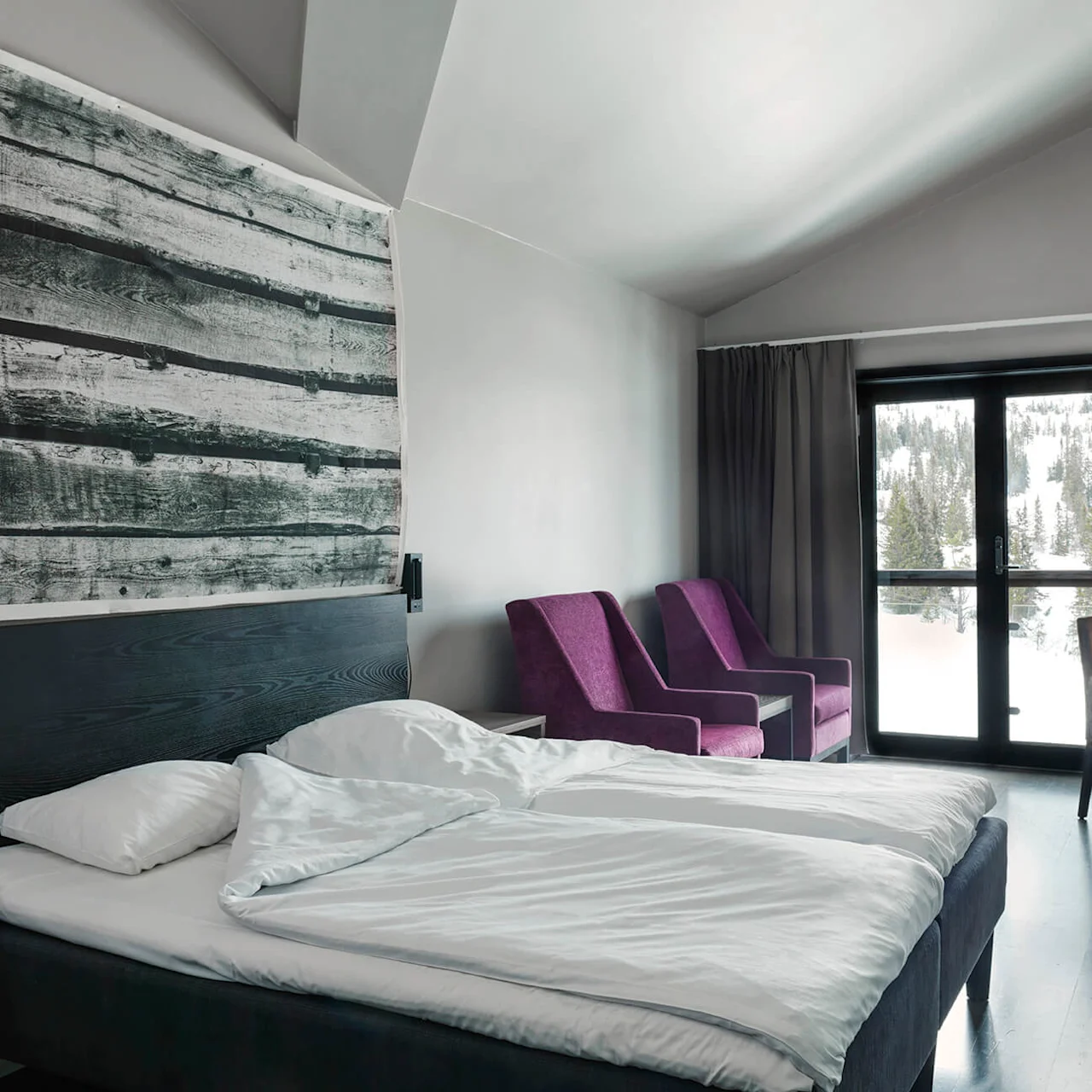 A bed with white linens sits in a modern room, accompanied by purple chairs and a desk with a lamp. Large windows reveal a snowy forest outside.
