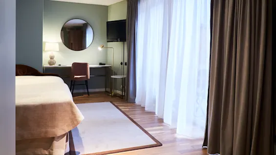 A modern hotel room with a plush bed, a work desk with a round mirror, soft lighting, and floor-length curtains partially drawn, revealing a window.