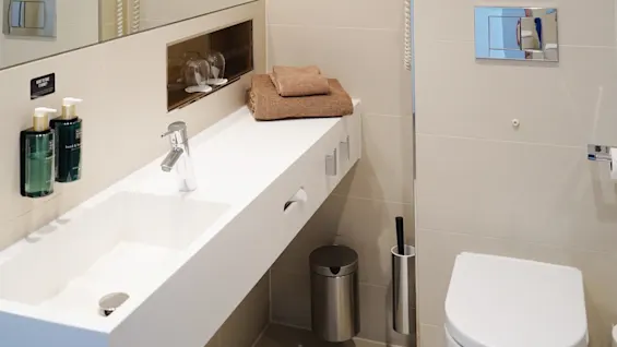 A modern bathroom features a white sink on a beige countertop with folded towels, under a large mirror. Wall-mounted toiletries and a hairdryer frame the space. Text: "HAND SOAP" and "BODY LOTION".