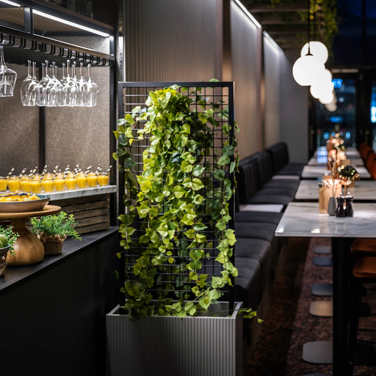 Fruit and drinks are arranged on a black countertop with glasses hanging overhead. A plant-covered partition separates the bar area from an elegantly set dining space with modern lighting.