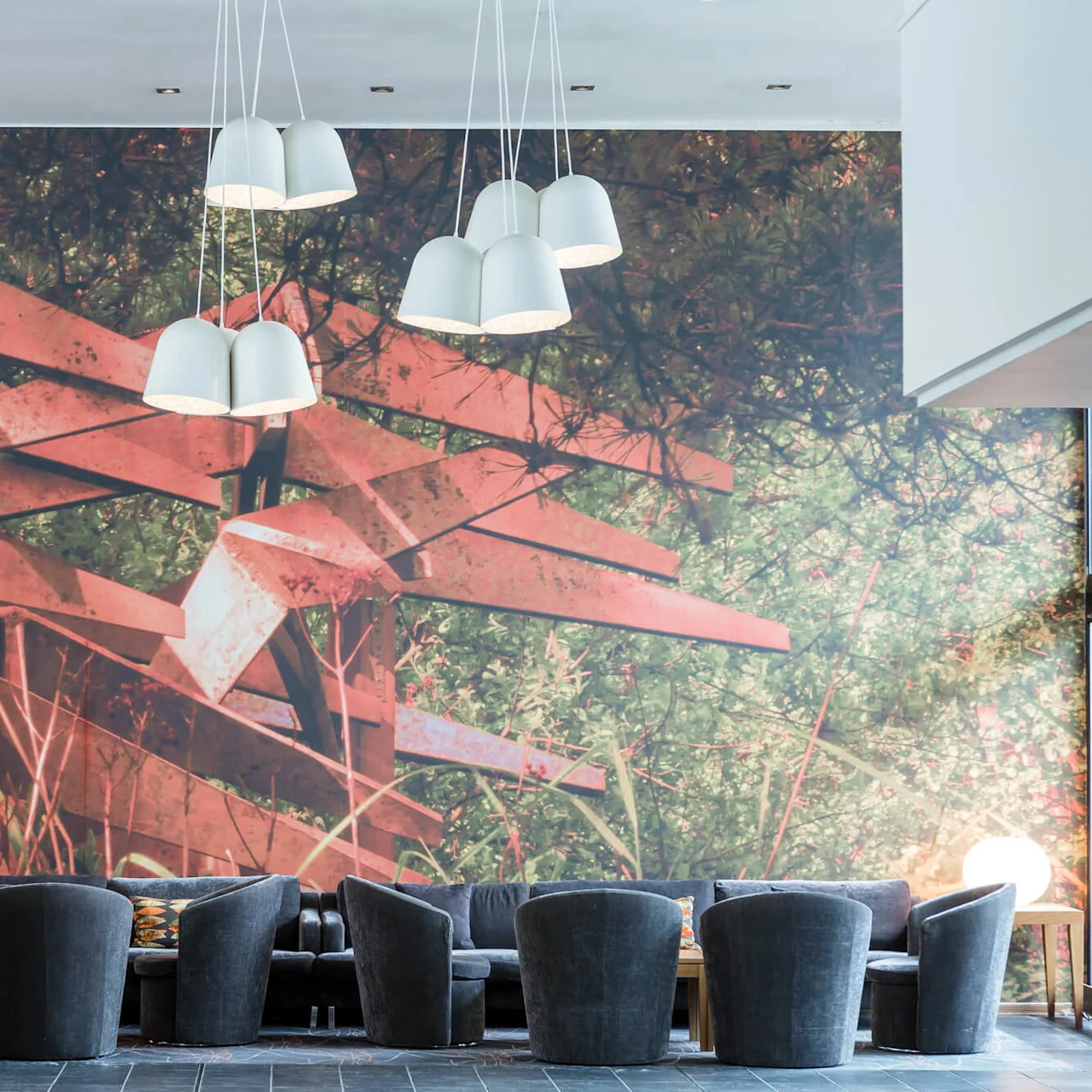 Gray armchairs form a seating arrangement in front of a wall-sized, abstract forest mural. Above, white pendant lamps hang from the ceiling. Natural light filters from the right.