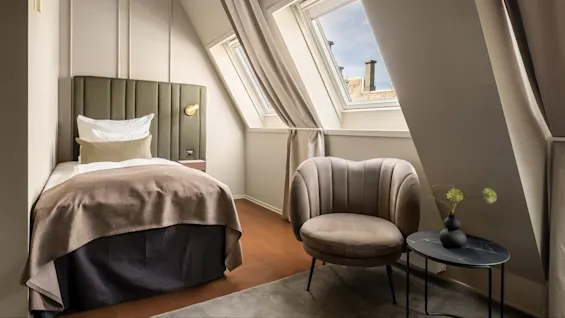 Et lille, moderne soveværelse har en seng med en grøn sengegavl og brunt sengetøj. Der er en blød stol og et lille bord med en vase ved siden af skråvinduer.