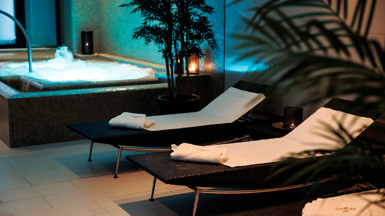 Loungers are aligned with towels on them, next to a bubbling hot tub. Ambient lighting and potted plants create a calming spa atmosphere.