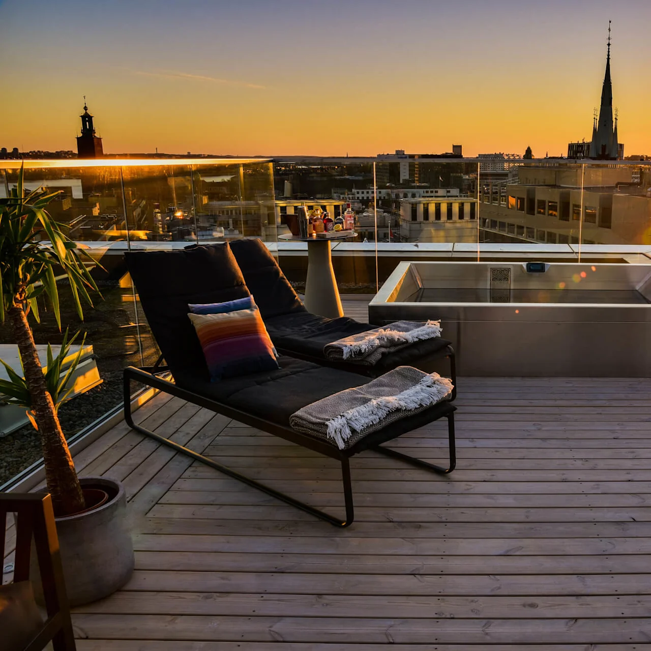 Takterrass-stoler med fargerike puter og tepper ser utover bysilhouetten, omgitt av glassrekkverk og bygninger opplyst av solnedgang. Planter og et lite bord kompletterer utemiljøet.