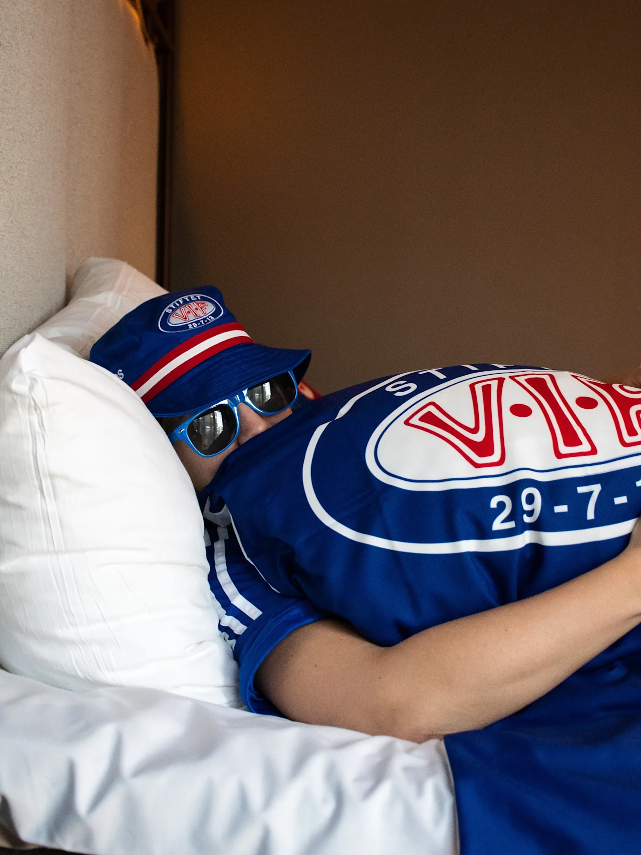 En person i blå hatt og solbriller slapper av i sengen, iført en blå skjorte. Begge plaggene har teksten VIF 29 7 13.