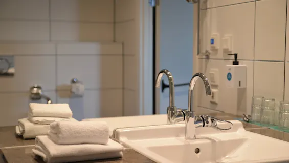 Vask med skinnende vandhaner står på en stenkonbordplade i et badeværelse. Hvide fliser på væggene, en bunke omhyggeligt foldede håndklæder og glas er i nærheden.