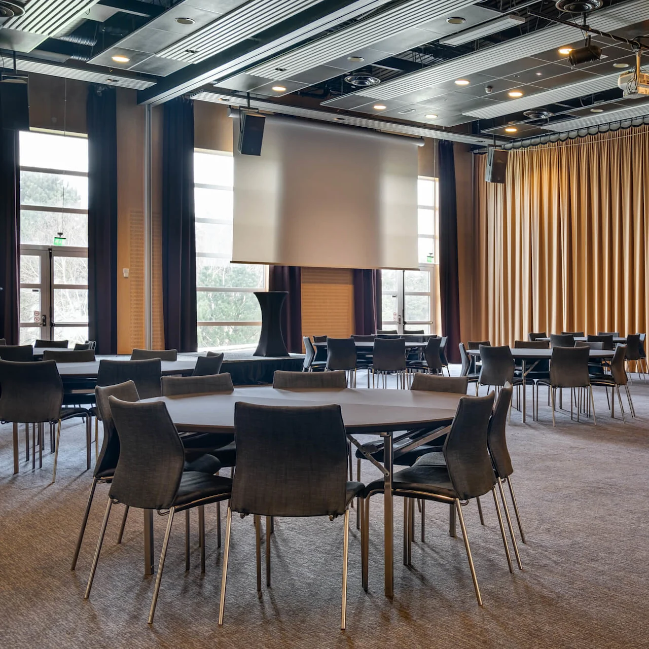 Empty conference room with round tables and chairs, large screen at the front, and long curtains on the side.